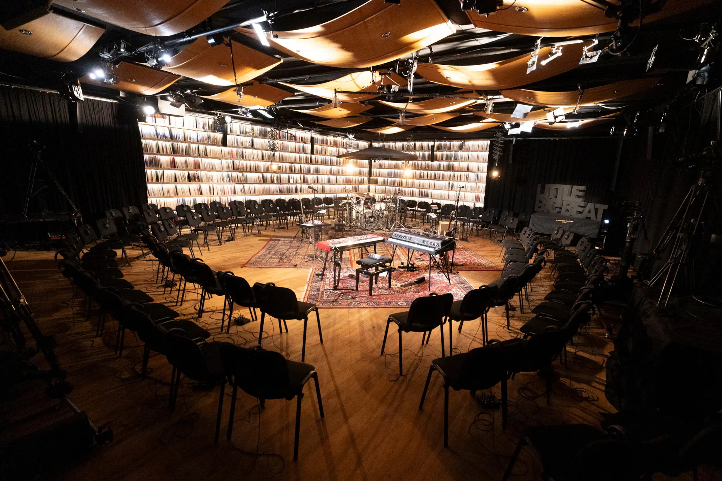 a performance room with a curved wooden ceiling, a wall of records, and chairs arranged in a circle around keyboards and drums