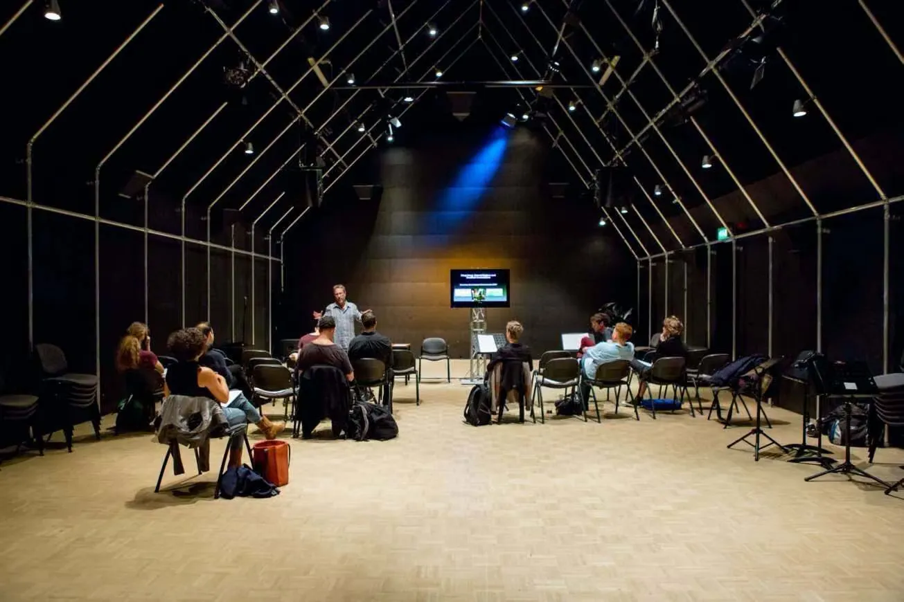 a hall with a pitched black roof, with a small group seated on chairs on a wooden floor